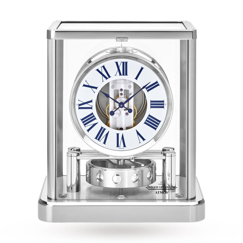 The Jaeger-LeCoultre Atmos Classique features a glass case, white dial with blue Roman numerals, visible internal mechanisms, and a sleek silver base for modern elegance.
