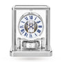 The Jaeger-LeCoultre Atmos Classique features a glass case, white dial with blue Roman numerals, visible internal mechanisms, and a sleek silver base for modern elegance.