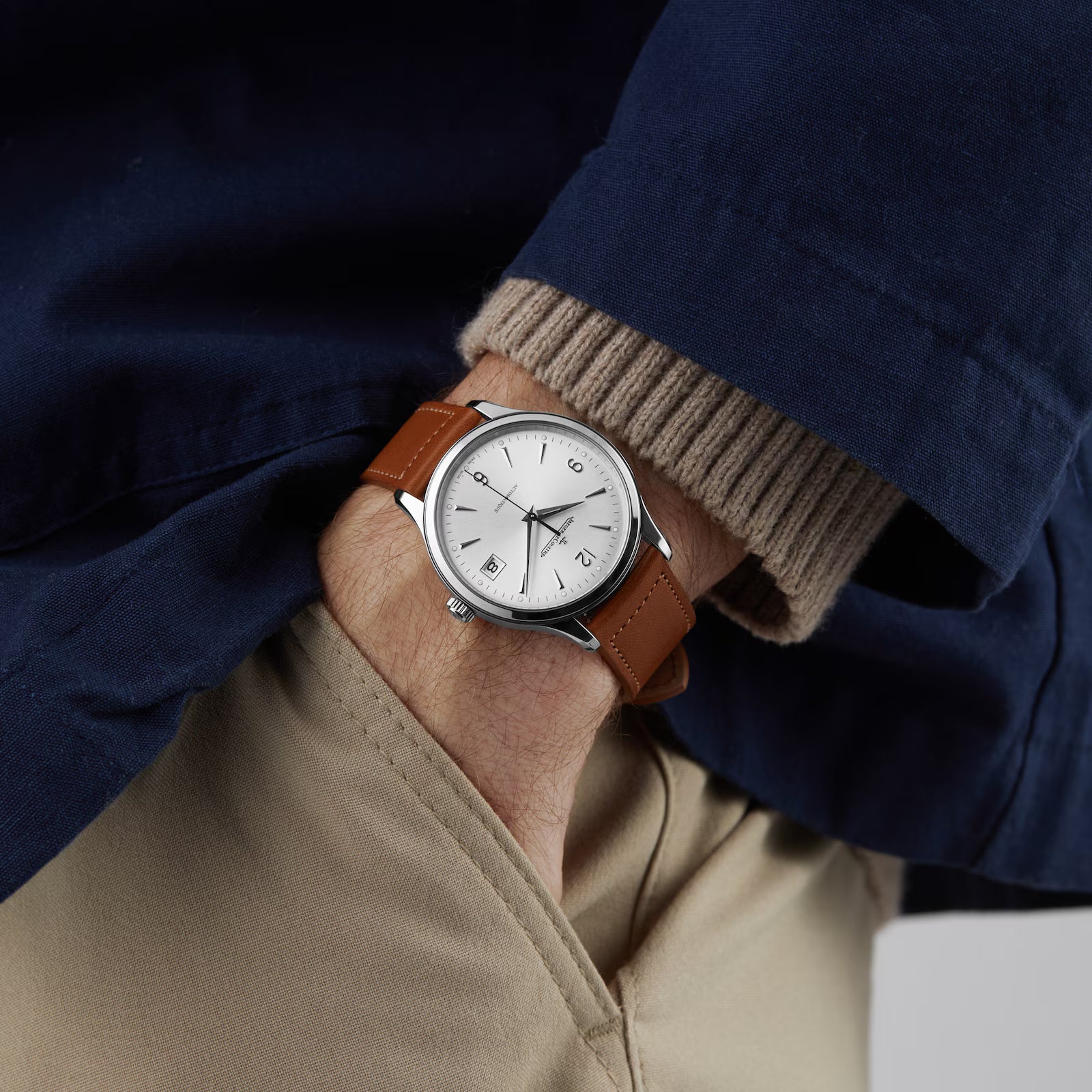A person in a navy jacket and beige pants shows off the Jaeger-LeCoultre Master Control Date watch, featuring a white dial, brown leather strap, and stainless steel case.