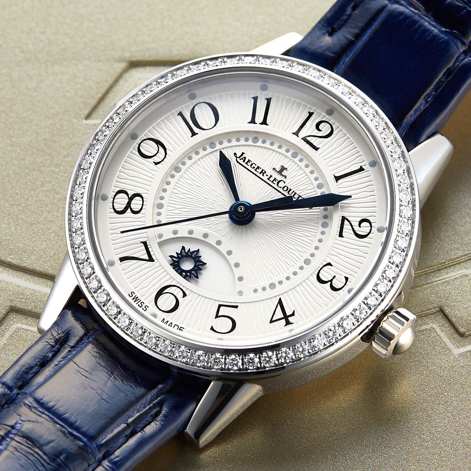 The Jaeger-LeCoultre Rendez-Vous Night & Day features a diamond-studded bezel, white textured dial with blue hands and Arabic numerals, sun and moon phase indicator, and a blue leather strap on a patterned surface.