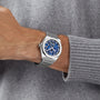 Someone wears the Zenith Defy Skyline Skeleton, a silver wristwatch with a blue skeleton dial exuding urban sophistication, paired with a gray knit sweater.