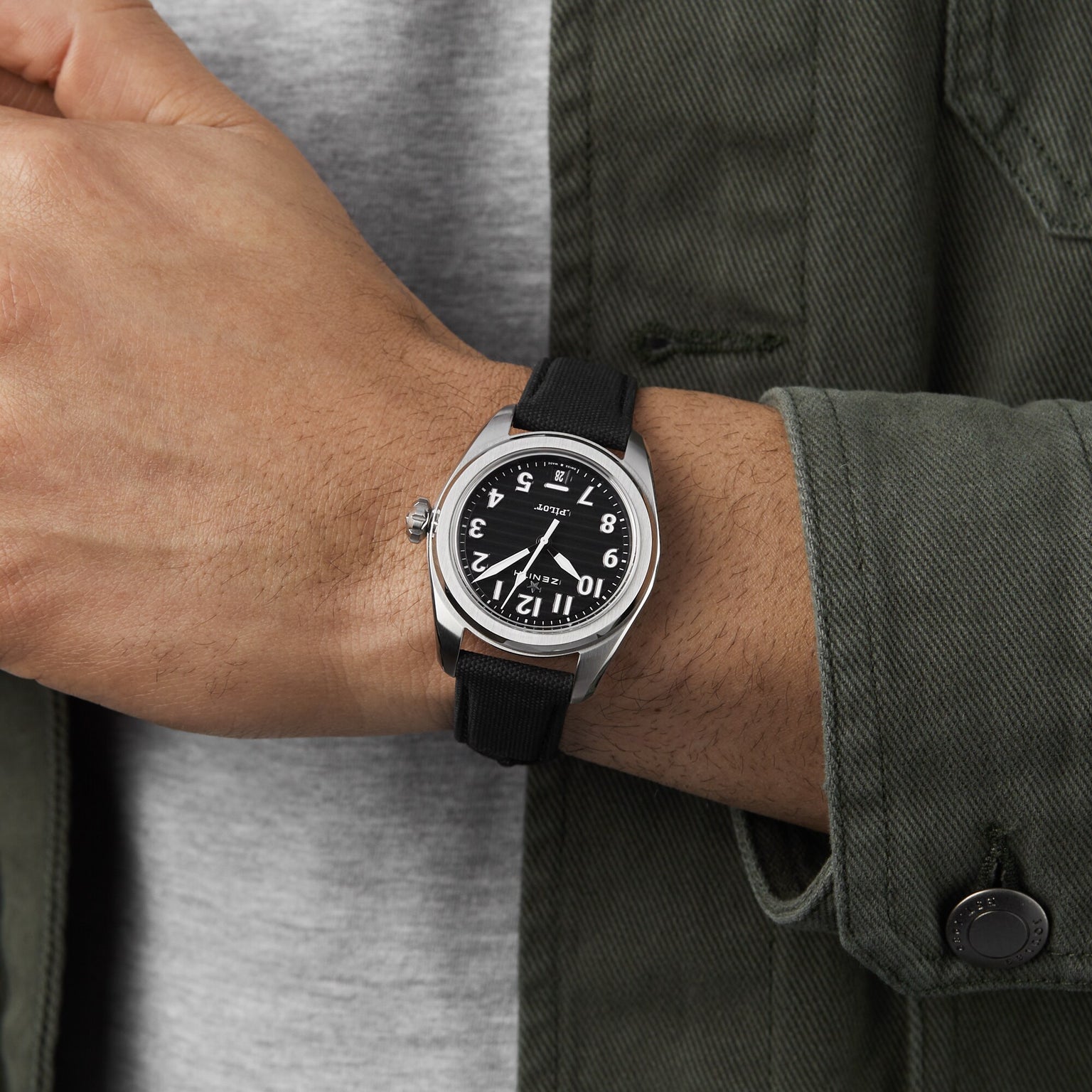 A person in a gray shirt and green jacket wears the Zenith Pilot Automatic watch, featuring a round black face, white numerals, black dial, and black strap.