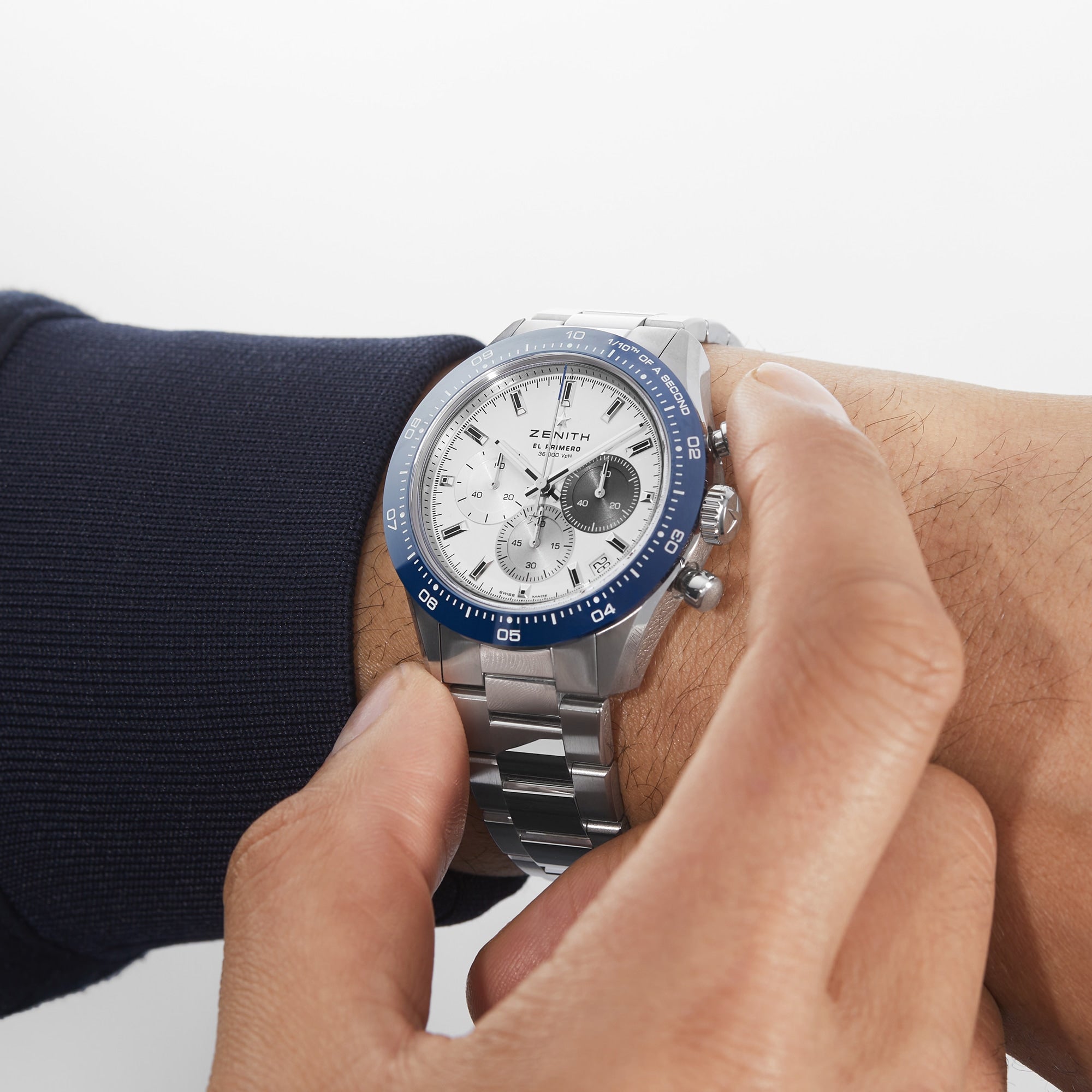 Someone adjusts the time on their left wrist, wearing a Zenith Chronomaster Sport Centenary Exclusive with a white face and blue bezel—an iconic ZENITH chronograph available at Watches of Switzerland.