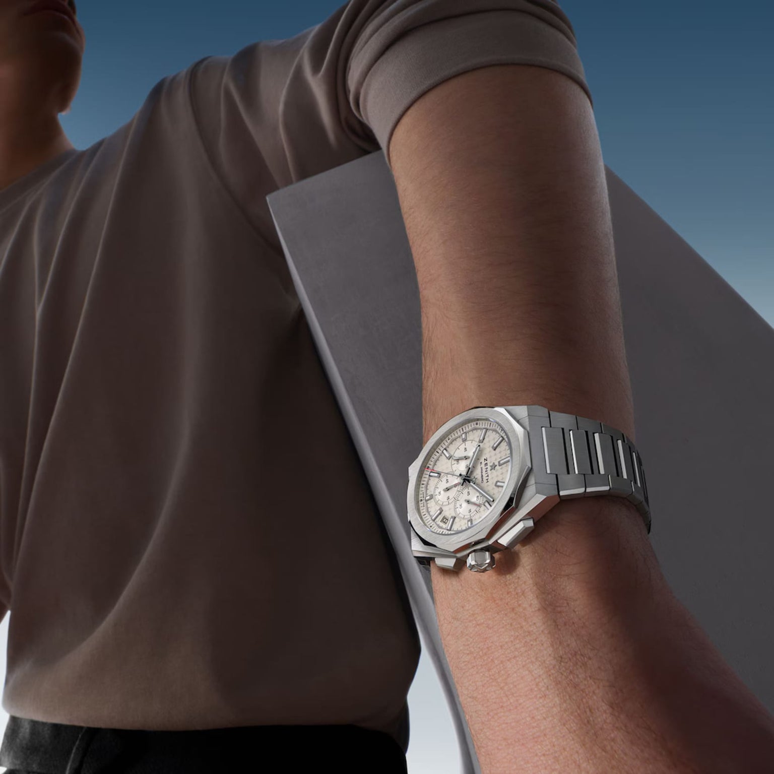 A person in a light beige shirt holds a rectangular gray object, wearing a Zenith Defy Skyline Chronograph with an interchangeable steel bracelet on their left wrist, set against a blue sky background.