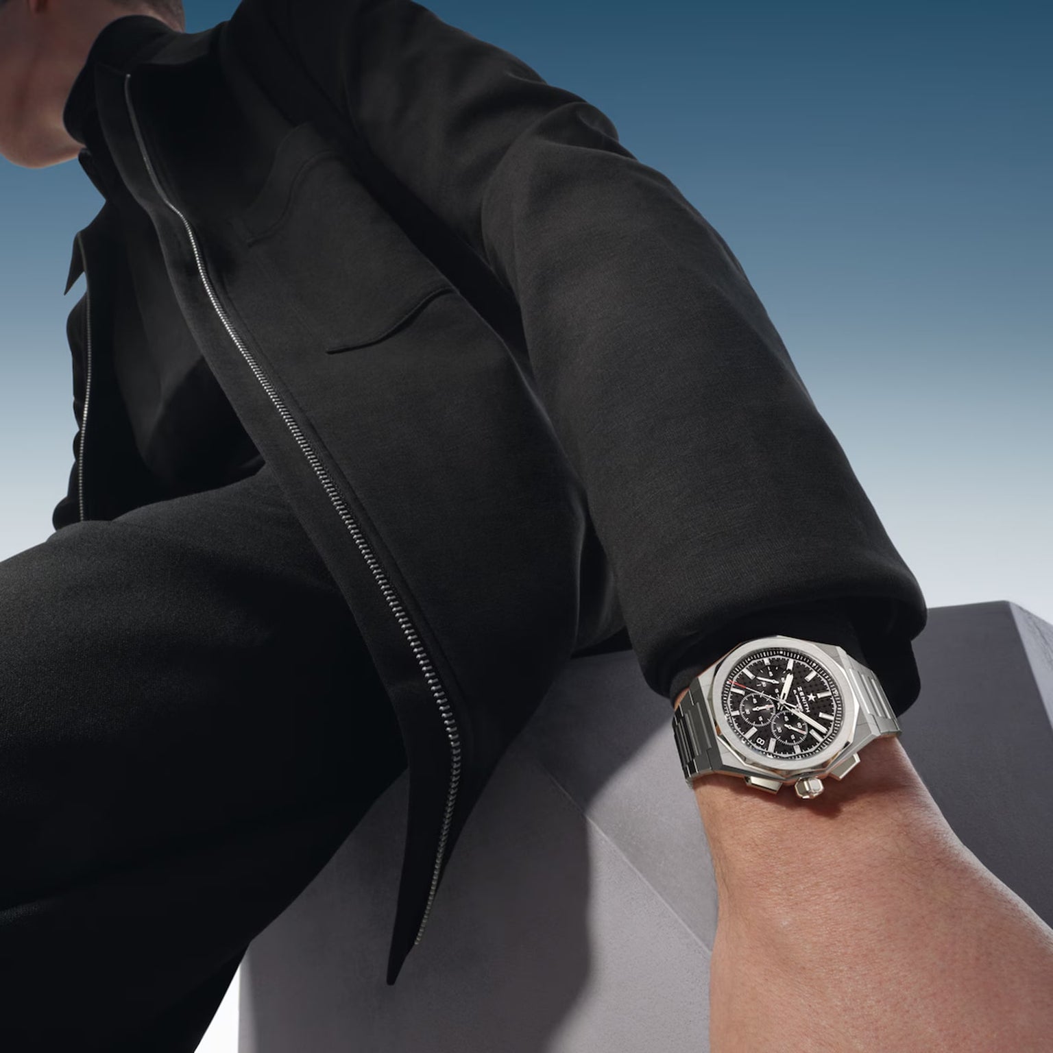 Wearing black, a person sits on a light surface, extending an arm to showcase the Zenith Defy Skyline Chronograph—a silver wristwatch with a black face.