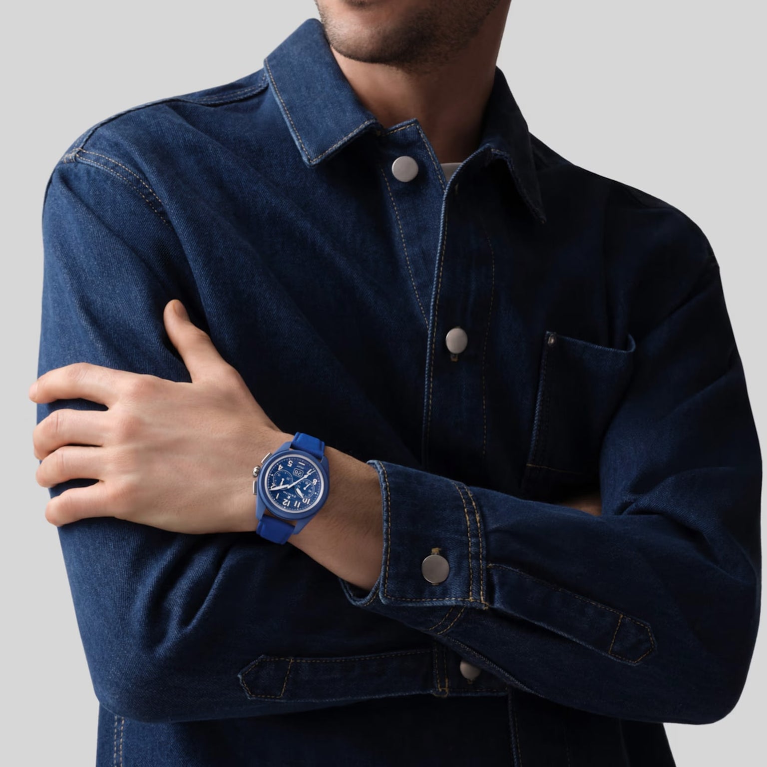 A person in a dark denim jacket crosses their arms, showing a Zenith Limited Edition Pilot Big Date Flyback 160th Anniversary Edition on their left wrist against a plain gray background.