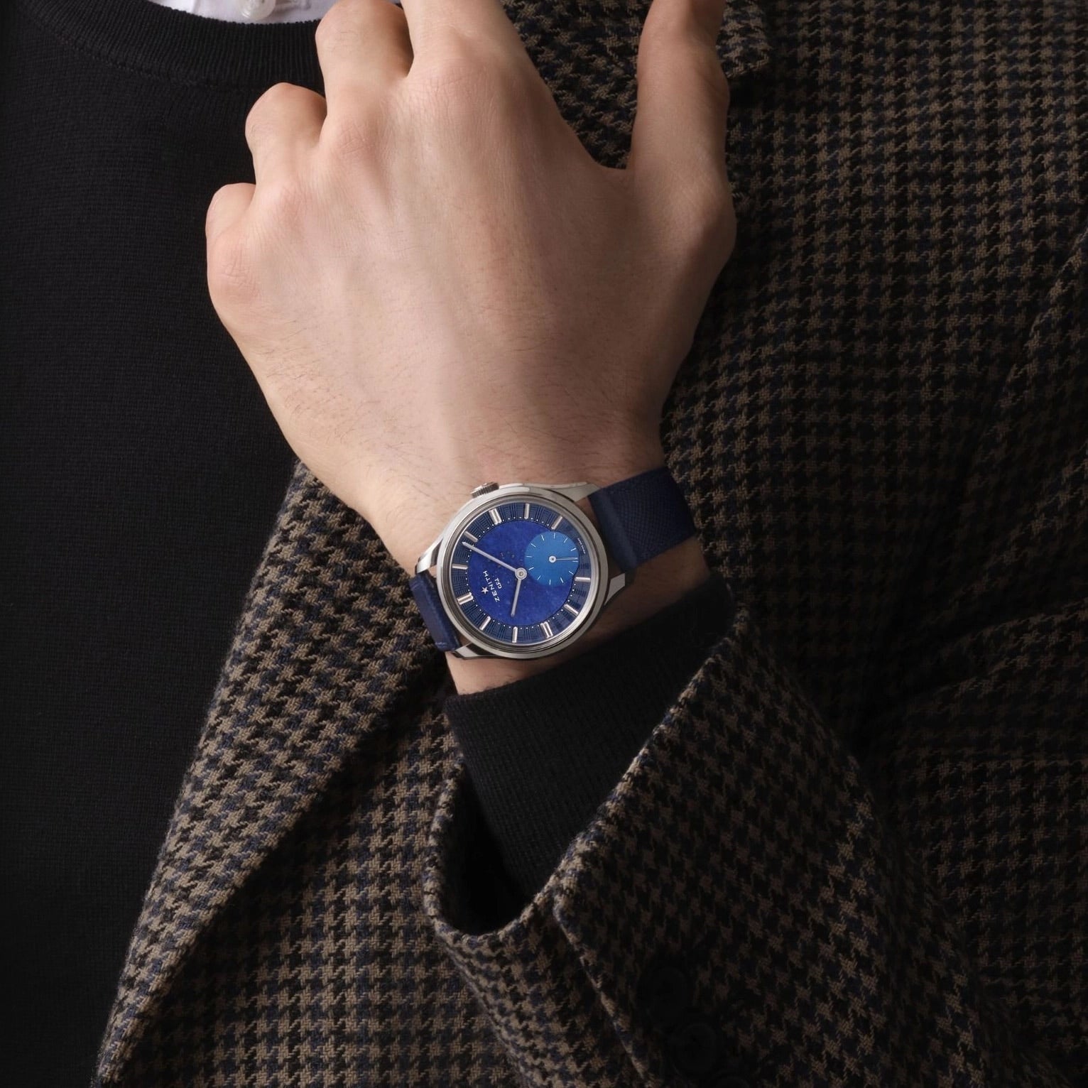 A person in a brown and black houndstooth jacket and black shirt showcases the Zenith Limited Edition G.F.J. Calibre 135 160th Anniversary Edition wristwatch with a platinum round case, blue dial, and blue strap.