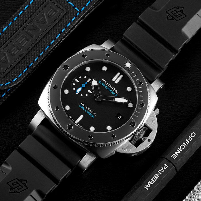 A close-up shows the Panerai Submersible men's dive watch with a black dial, 42mm stainless steel case, blue accents, and black rubber strap next to branded Panerai packaging and accessories.