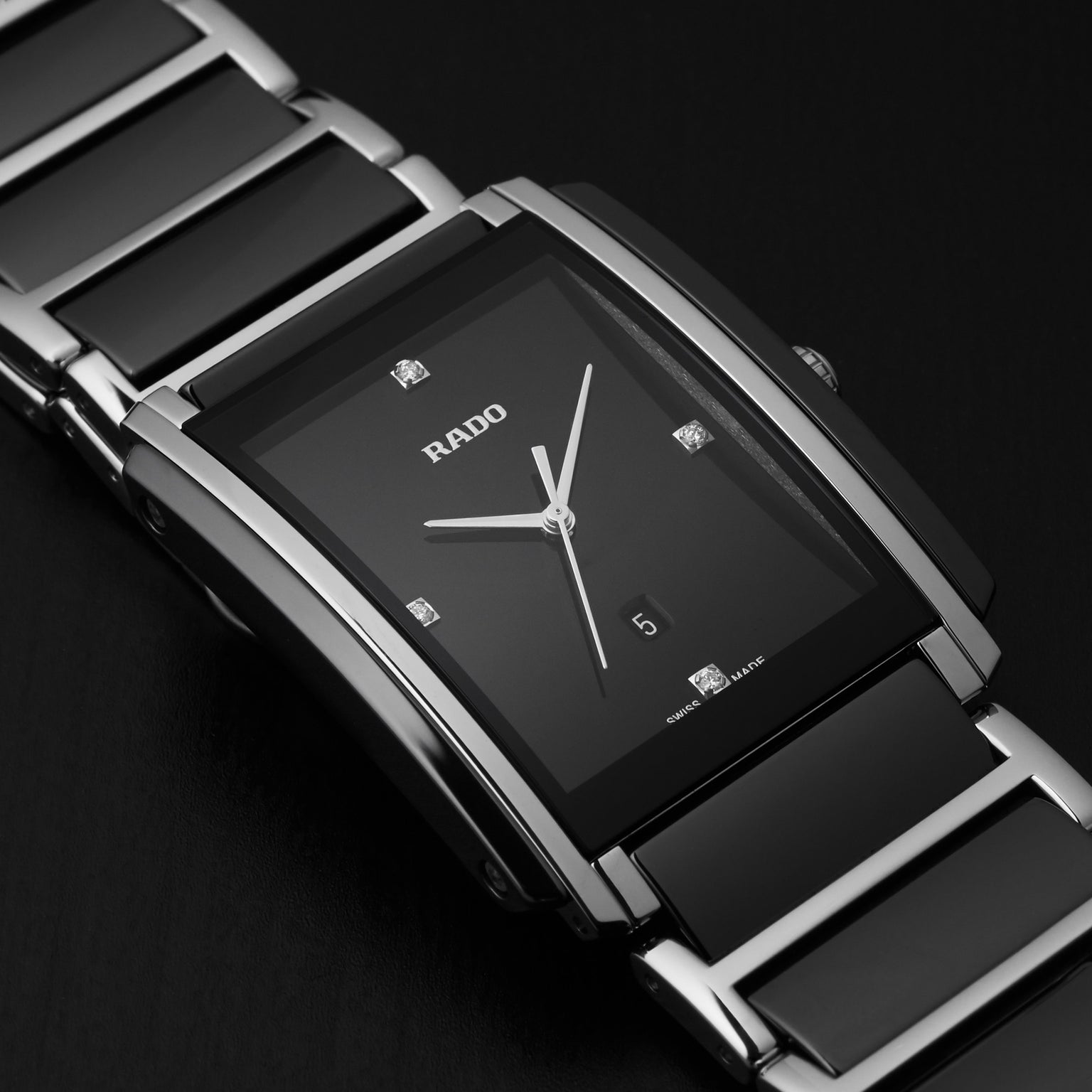 A close-up of the Rado Integral Diamonds watch showcases its rectangular black dial with silver hour markers, diamond accents, and a metal bracelet. Featuring high-tech ceramic and quartz movement, this timepiece stands out against a dark background.