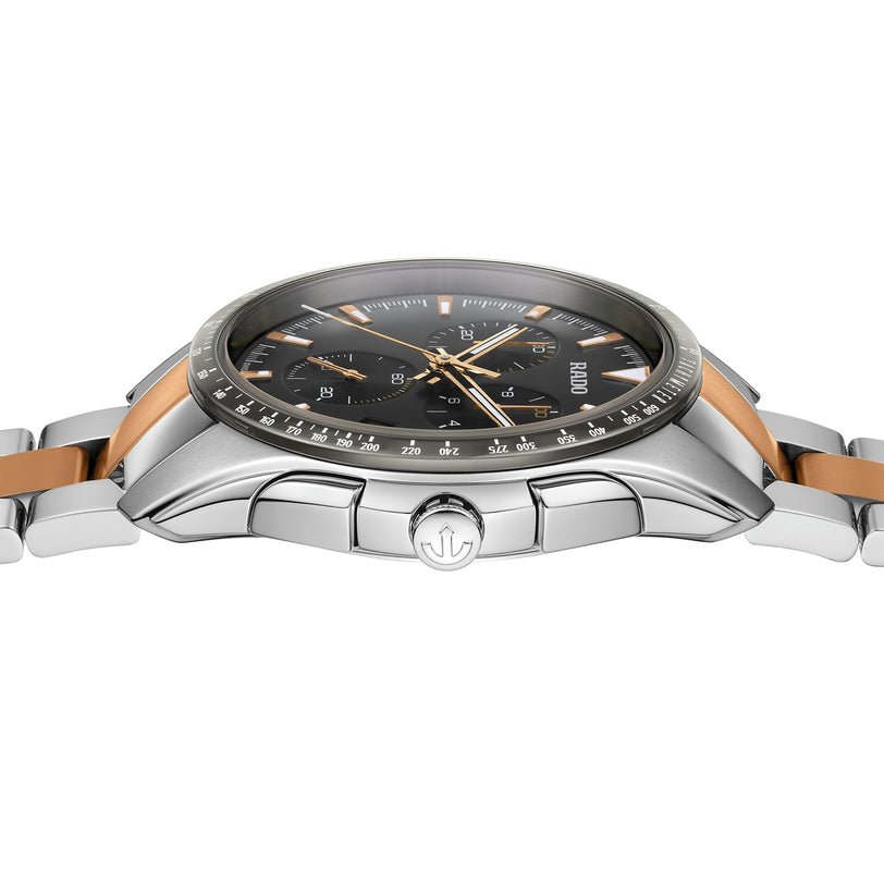 A close-up side view of the Rado Hyperchrome Chronograph, featuring a silver and rose gold case, black dial, metal bracelet, and signature high-tech ceramic design with detailed crown and case elements.