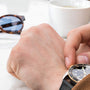 A person checks the time on a TUDOR Black Bay S&G wristwatch while sitting at a table with sunglasses and a cup of coffee in the background.