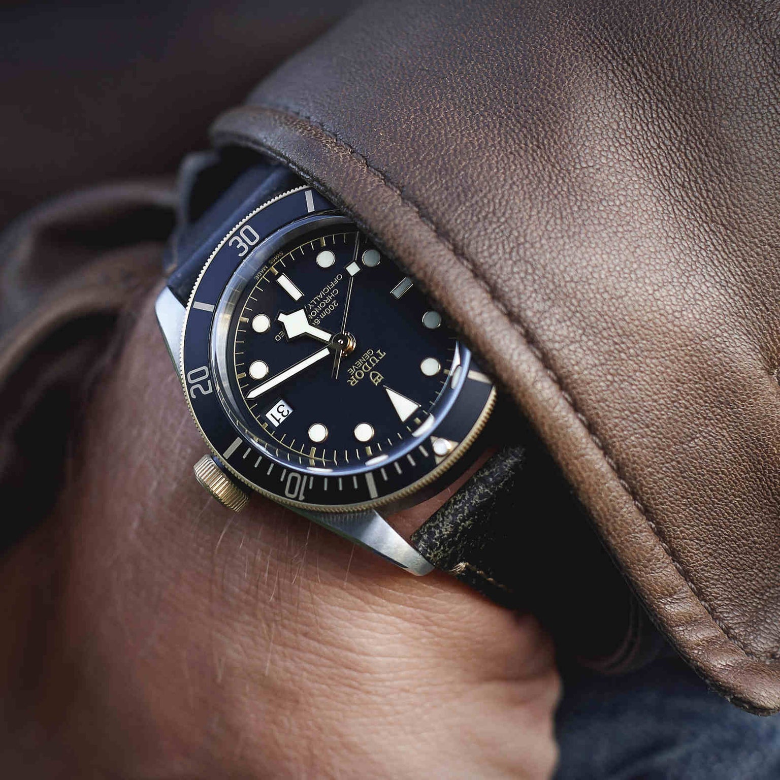 A wrist sporting the TUDOR Black Bay S&G dive watch, known for its timeless style and distinguished by a black bezel and dial with gold accents, paired with a leather strap and partially covered by a brown leather jacket.