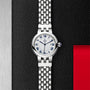 The TUDOR Clair de Rose features a silver linked bracelet, white dial, and blue Roman numerals set against a textured black background.