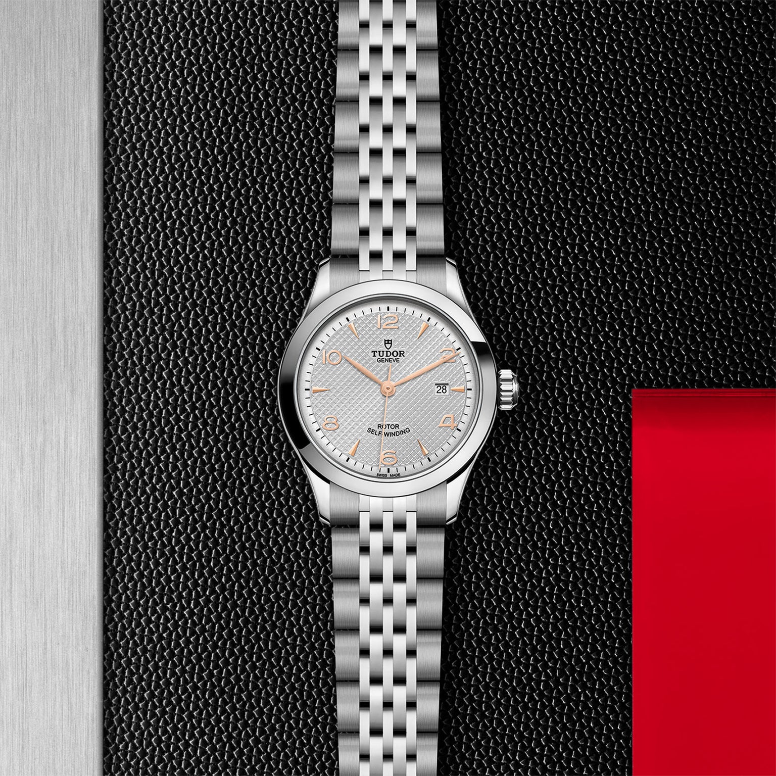 A TUDOR 1926 ladies’ wristwatch with a linked silver band and textured white dial is displayed on a black textured surface featuring metal and red accents.