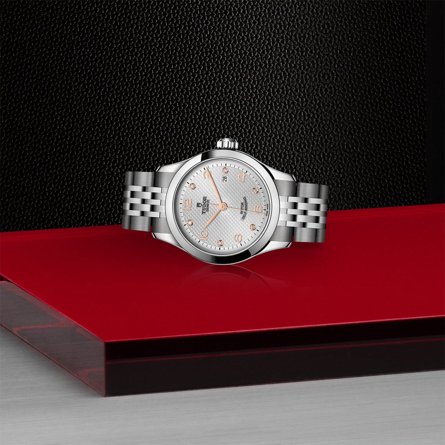A TUDOR 1926 wristwatch with a silver-tone metal band and white textured dial rests on a glossy red and gray surface, set against a black background, highlighting the refined elegance of mechanical timepieces.