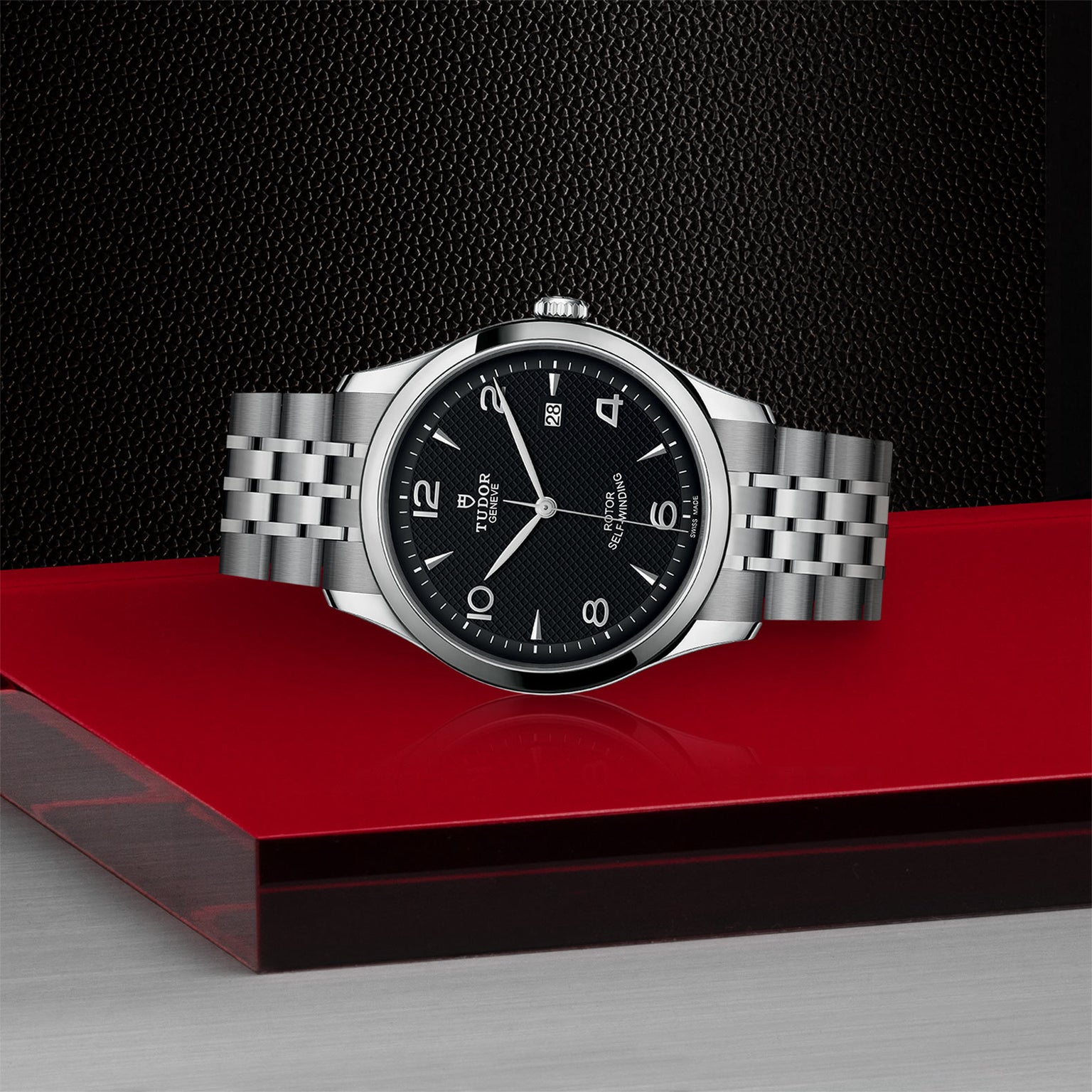 The TUDOR 1926, a silver wristwatch with a black dial and metal band, is displayed on a red and gray surface.