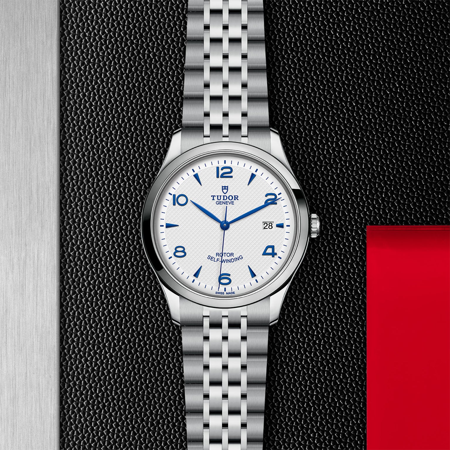 The TUDOR 1926 men’s watch pairs a silver metal bracelet with a white dial and blue Arabic numerals, elegantly displayed on a textured black and red background—an ideal pick for mechanical watch enthusiasts.