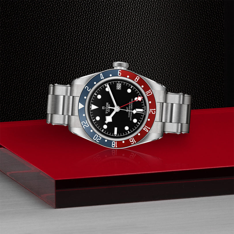 The TUDOR Black Bay GMT is a stainless steel wristwatch with a red and blue bezel, black dial, and metal bracelet displayed on a red surface.