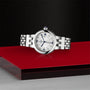 The TUDOR Clair de Rose, a silver luxury wristwatch with a white face, is displayed on a red and silver surface against a black background.