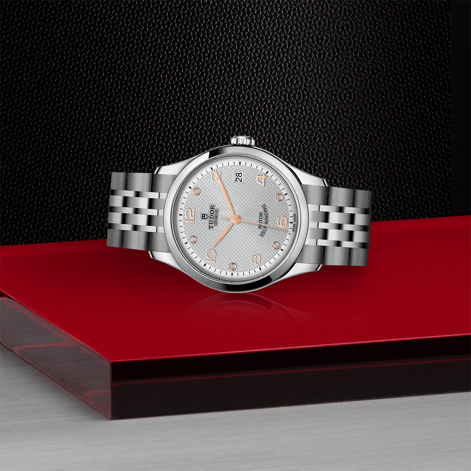 A TUDOR 1926 silver wristwatch with a metal band is displayed on a red surface, set against a textured black background.