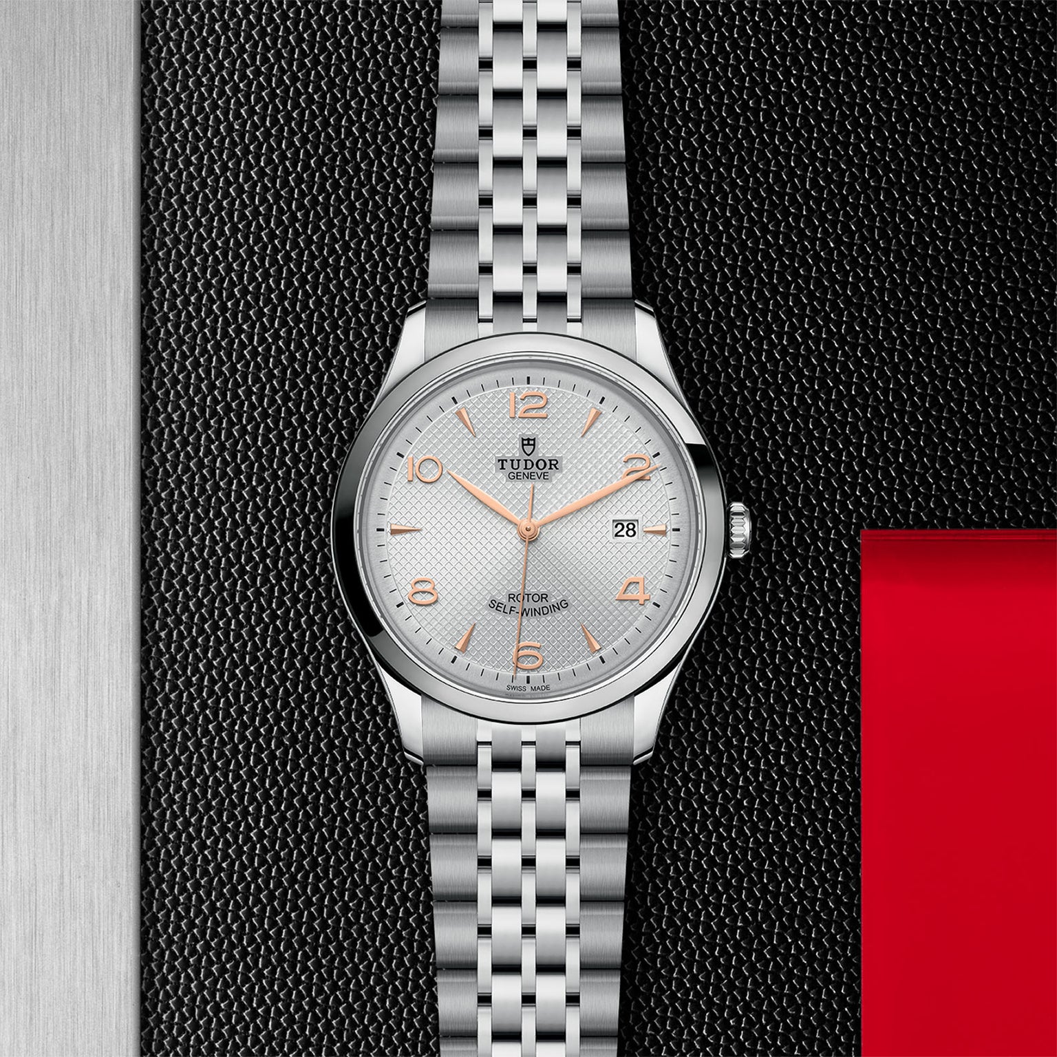 A TUDOR 1926 silver wristwatch with an orange-accented dial, self-winding movement, and a metal link band is showcased on a textured black surface with red and silver panels.