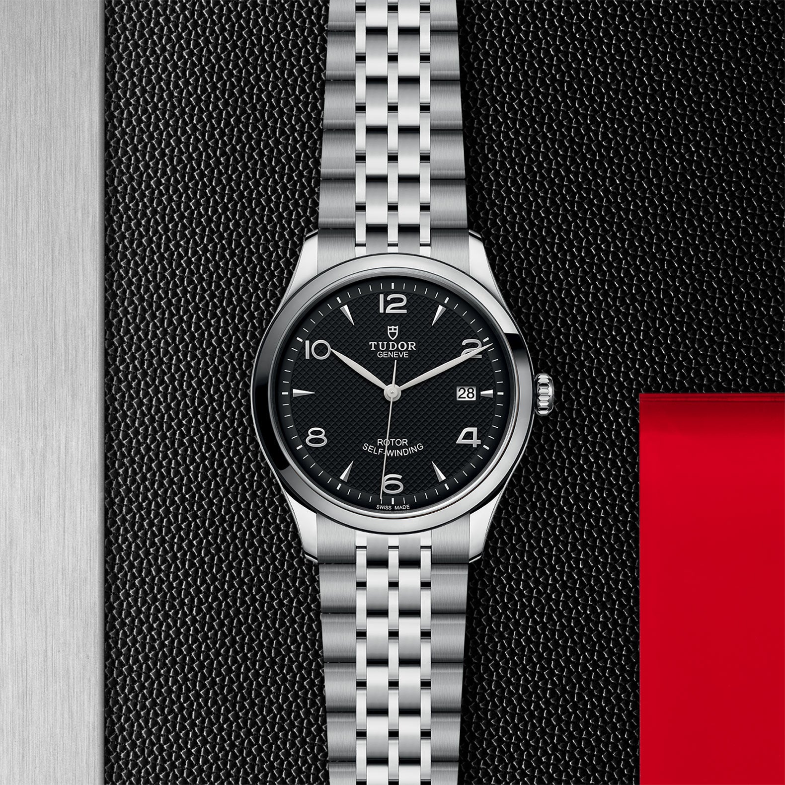 The TUDOR 1926 watch features a silver case, black dial, date display, and stainless steel bracelet, showcased on a textured black and metallic surface with red accents to highlight the brands signature elegance.