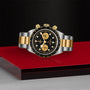 The TUDOR Black Bay Chrono S&G, a two-tone stainless steel and gold watch with a black and gold dial and Swiss-made movement, is displayed on a red and silver surface against a textured black background.