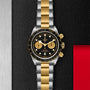 The TUDOR Black Bay Chrono S&G, featuring a black and gold dial, snowflake hands, gold bezel, and a gold and silver bracelet, is displayed on a textured black background.