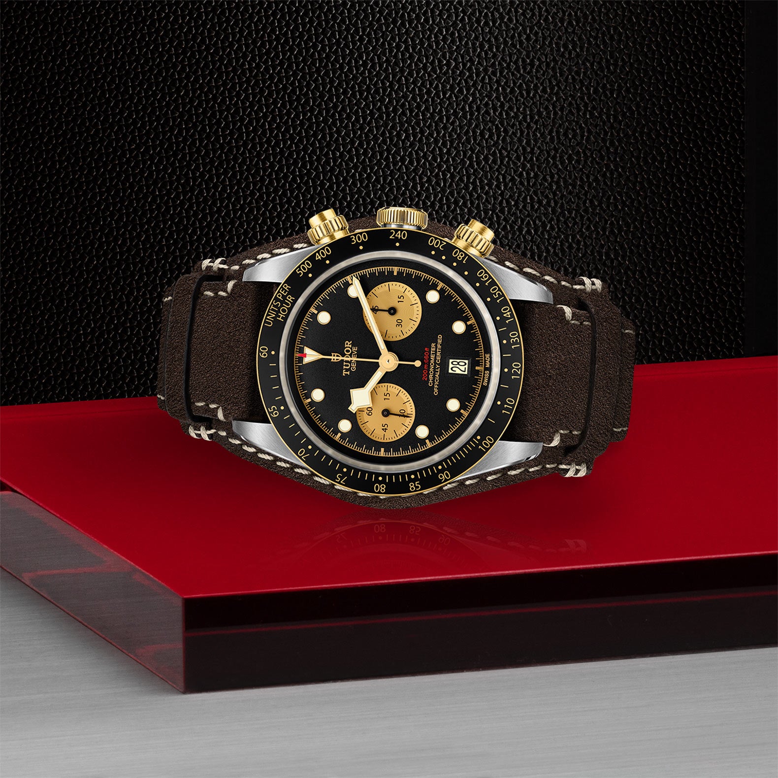 The TUDOR Black Bay Chrono S&G wristwatch with a brown leather strap is showcased on a red and clear platform against a textured black background, highlighting the signature design elements of the brand.