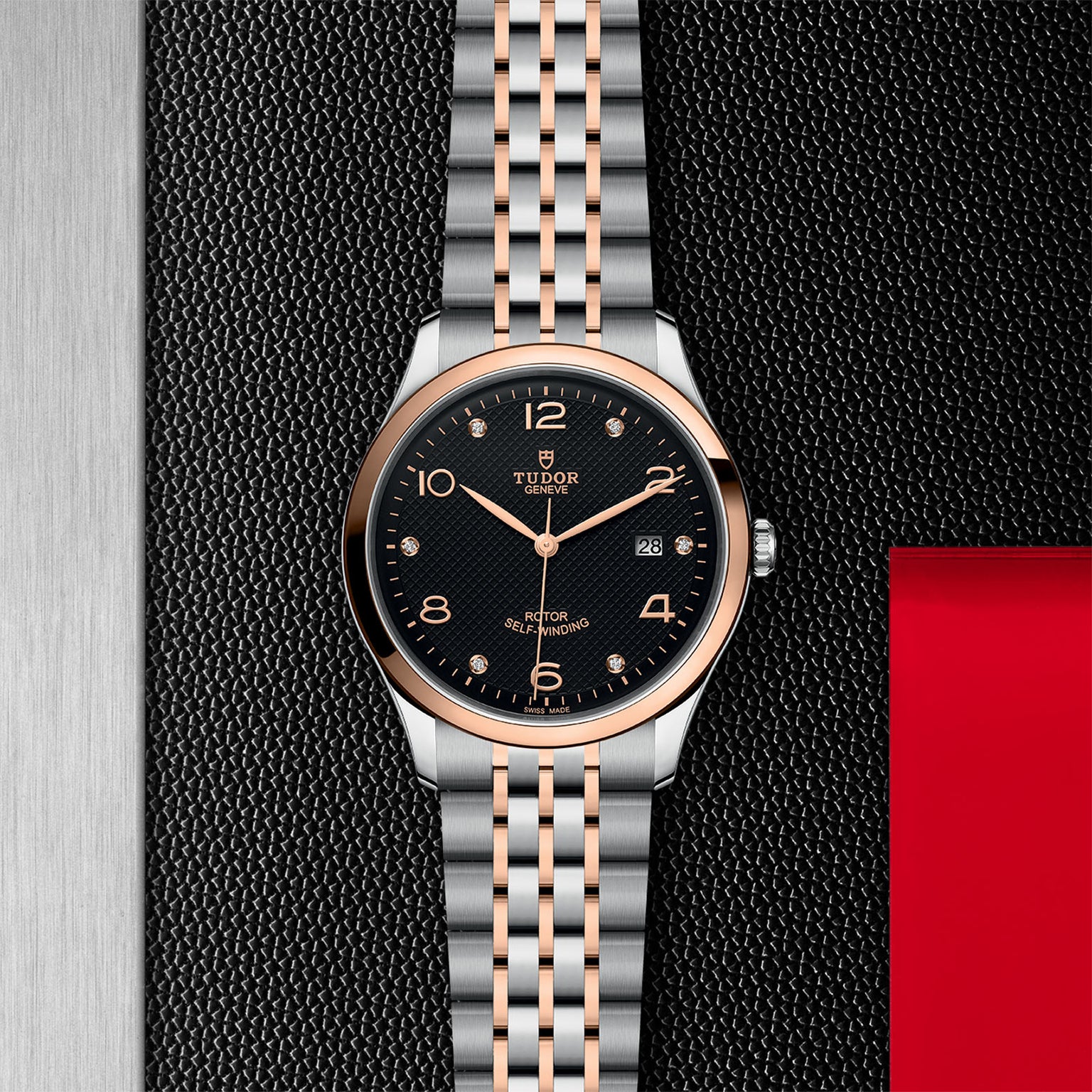 A TUDOR 1926 wristwatch with a black dial, gold accents, and a two-tone silver and gold bracelet is set against a textured black and silver background with red, showcasing the timeless elegance of mechanical design.