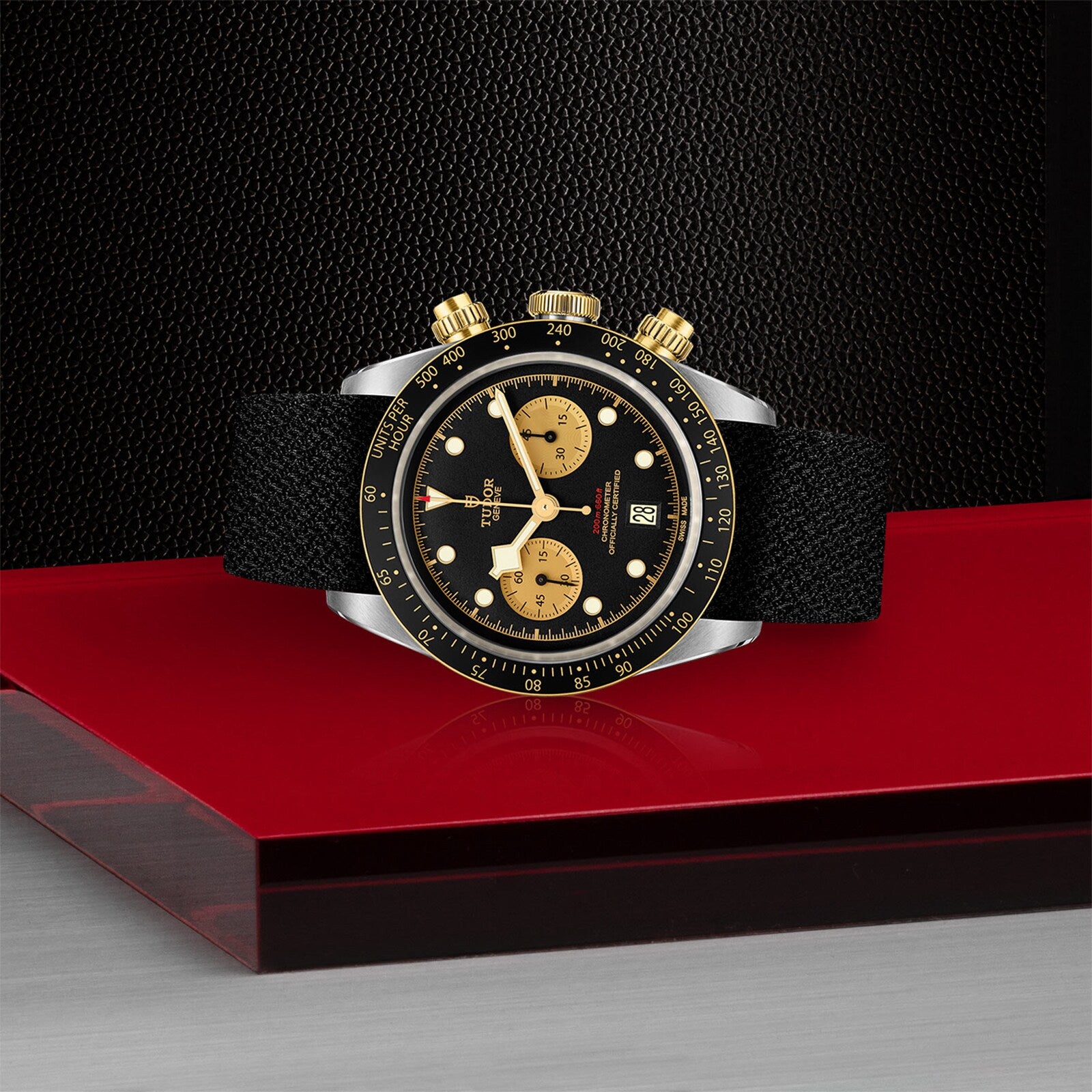 The TUDOR Black Bay Chrono S&G, a black and gold chronograph inspired by classic divers’ watches with a black strap, is displayed on a red and silver platform against a textured black background.