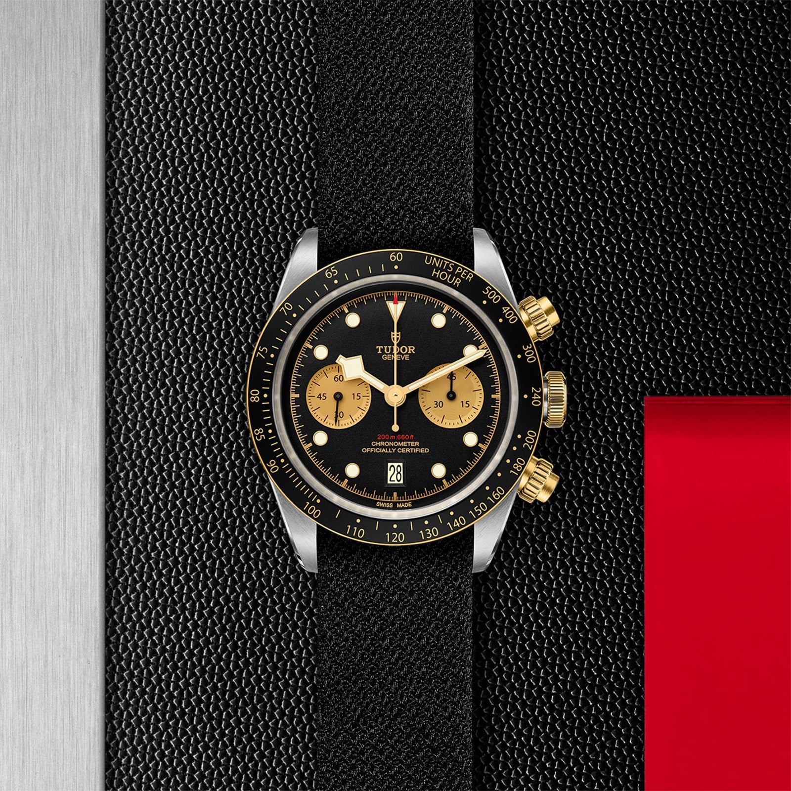 The TUDOR Black Bay Chrono S&G, featuring a black and gold chronograph dial and a black strap, is displayed on a textured black surface with metallic and red accents.