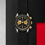 The TUDOR Black Bay Chrono S&G, featuring a black and gold chronograph dial and a black strap, is displayed on a textured black surface with metallic and red accents.