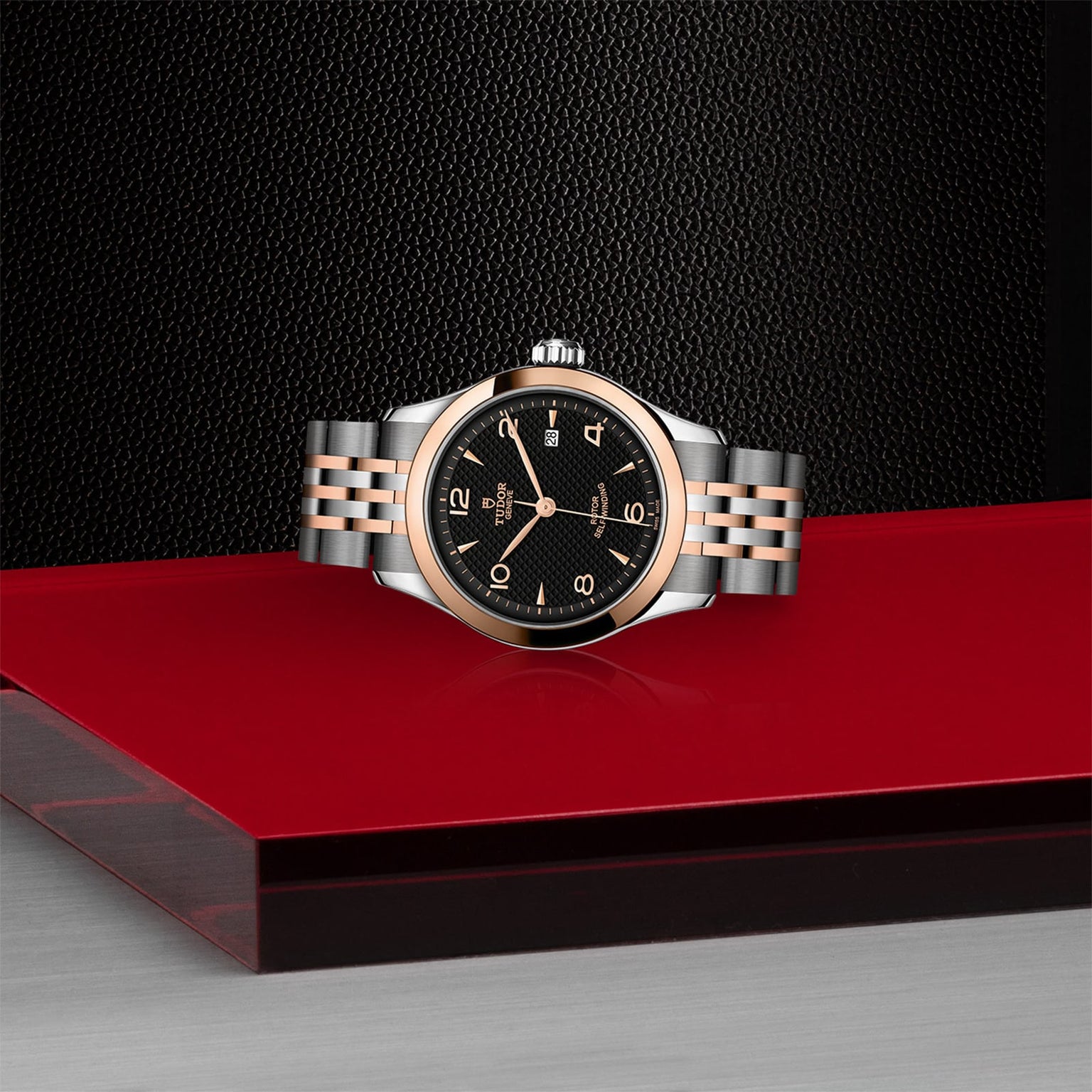 The TUDOR 1926, a classic two-tone wristwatch with a black dial and silver and rose gold bracelet, is displayed on a glossy red and black surface with a textured background, highlighting the elegance of mechanical watches.