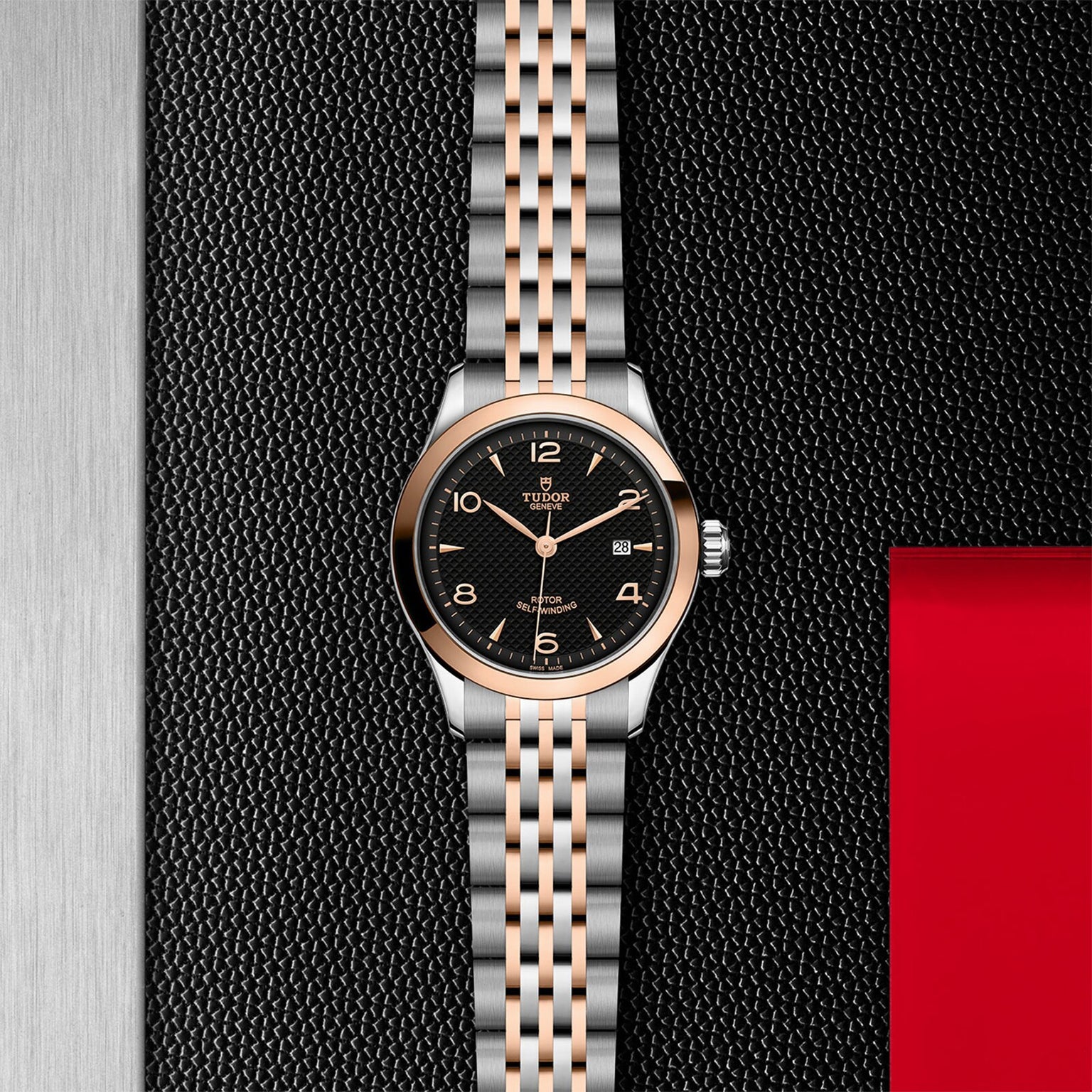 The TUDOR 1926 is a classic two-tone wristwatch featuring a black textured dial, gold hour markers, and date window, showcased against a black, silver, and red textured background.