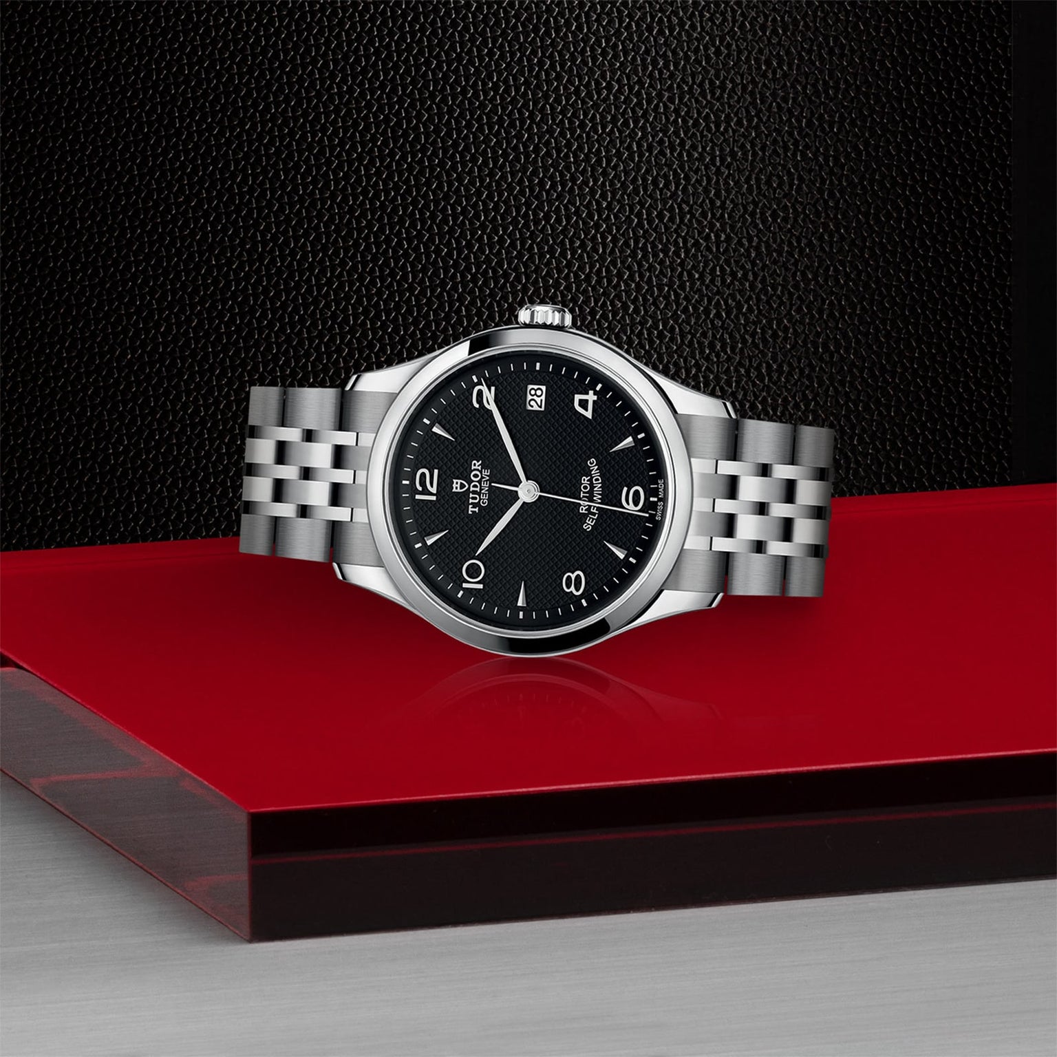 The TUDOR 1926, a classic stainless steel wristwatch with a black dial and silver bracelet, is showcased on a red and black platform against a textured dark background.