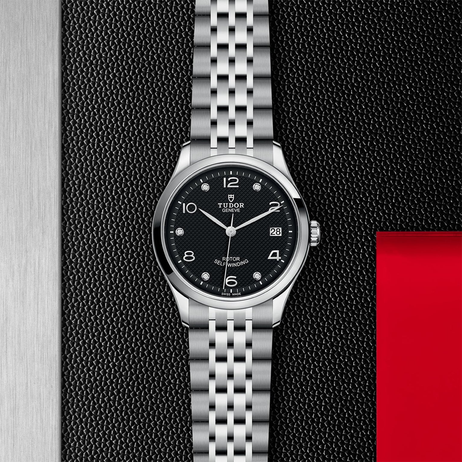 The TUDOR 1926 wristwatch, with a black dial, silver hour markers, and date window, rests on a dark textured surface with a red accent—a stylish nod to TUDOR’s enduring heritage.