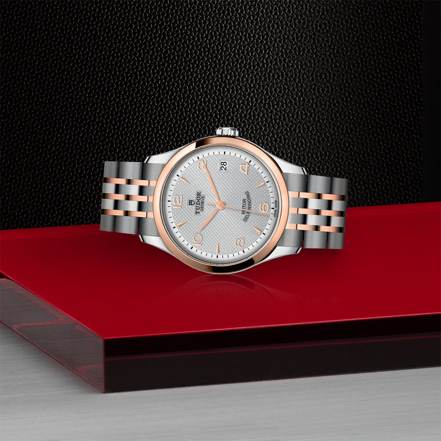 The TUDOR 1926 wristwatch in silver and rose gold, with a white textured dial, is displayed on a red surface against a black backdrop, highlighting the classic elegance of mechanical timepieces.