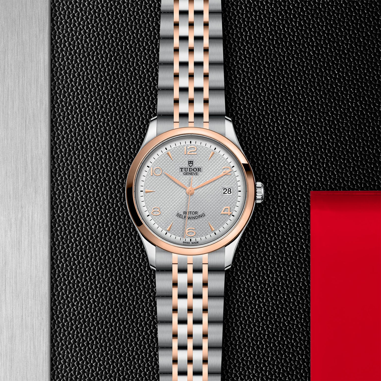 The TUDOR 1926 wristwatch features a silver and rose gold bracelet, silver dial, rose gold markers and hands, plus a date display—presented on a textured black and red background for classic watch enthusiasts.