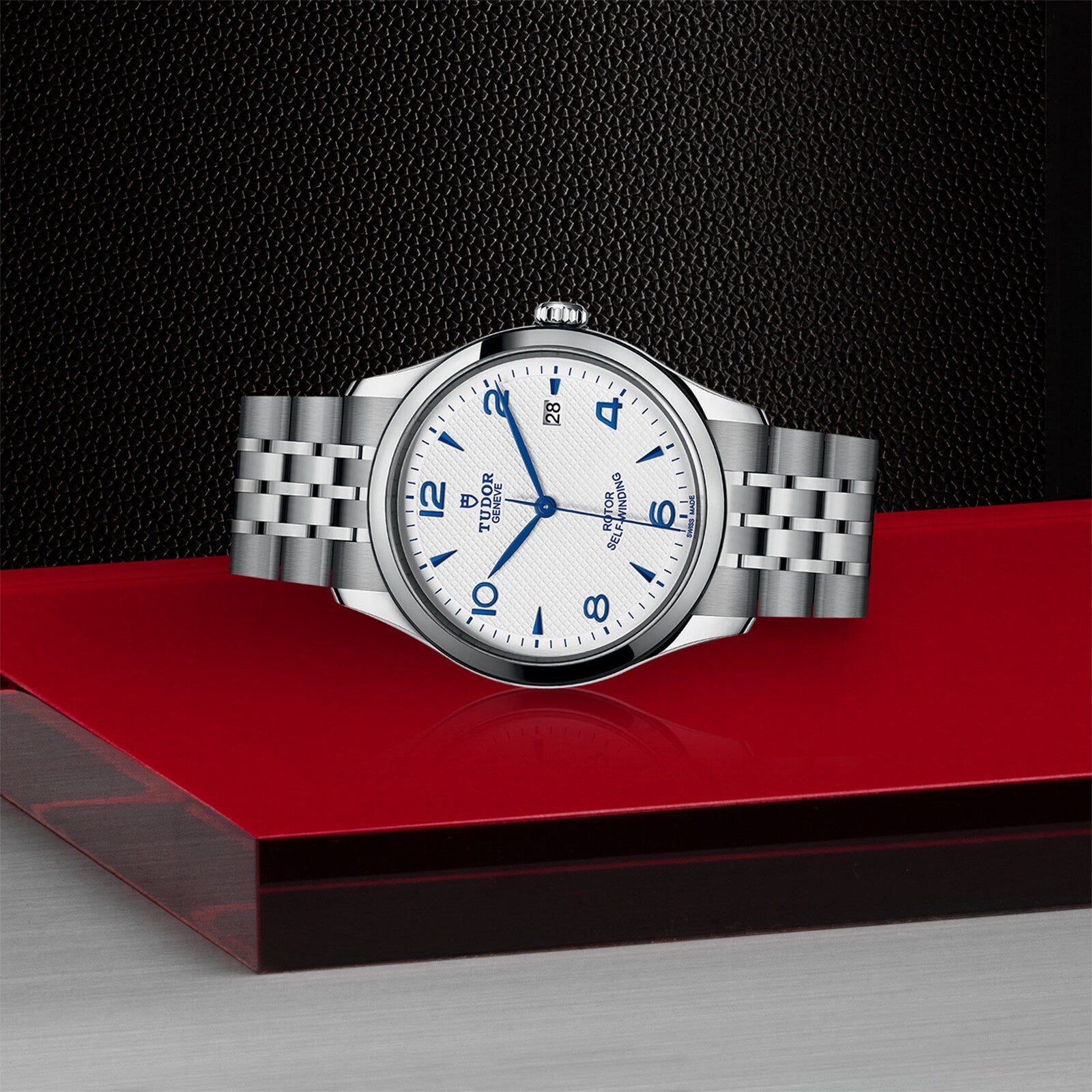 A TUDOR 1926 stainless steel wristwatch with a white dial, blue hour markers, and a metal bracelet is displayed on a glossy red and black surface.