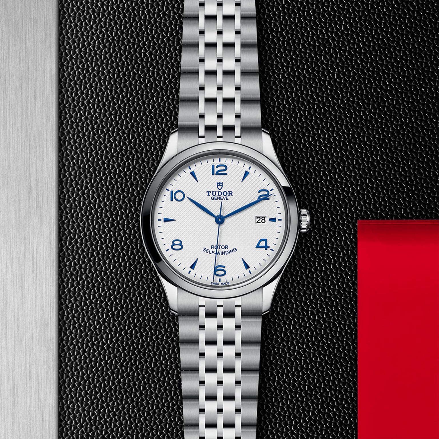 A TUDOR 1926 silver wristwatch featuring a white dial with blue numerals, date display, and silver metal bracelet, shown on a textured black background with red and silver accents.