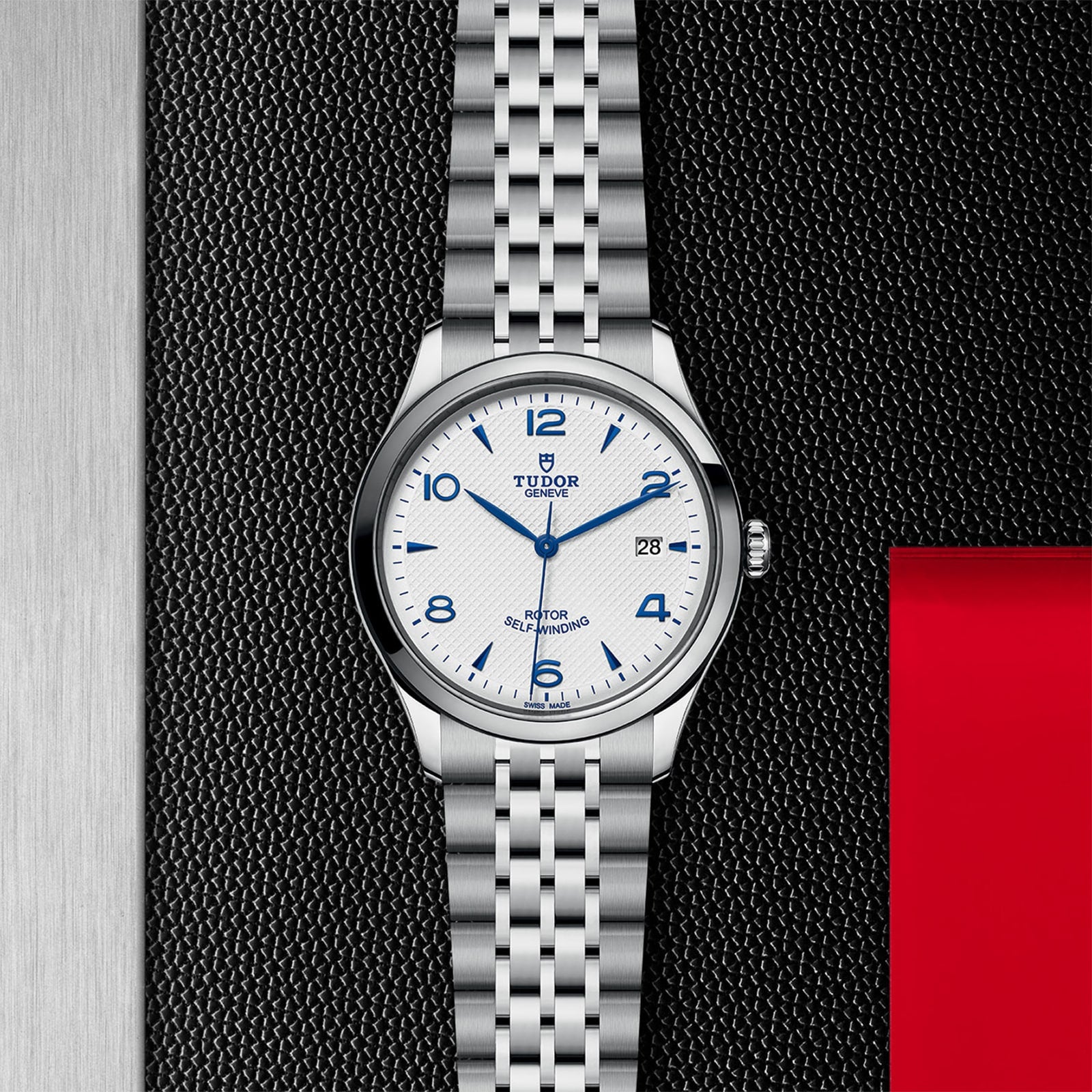 A TUDOR 1926 silver wristwatch featuring a white dial with blue numerals, date display, and silver metal bracelet, shown on a textured black background with red and silver accents.