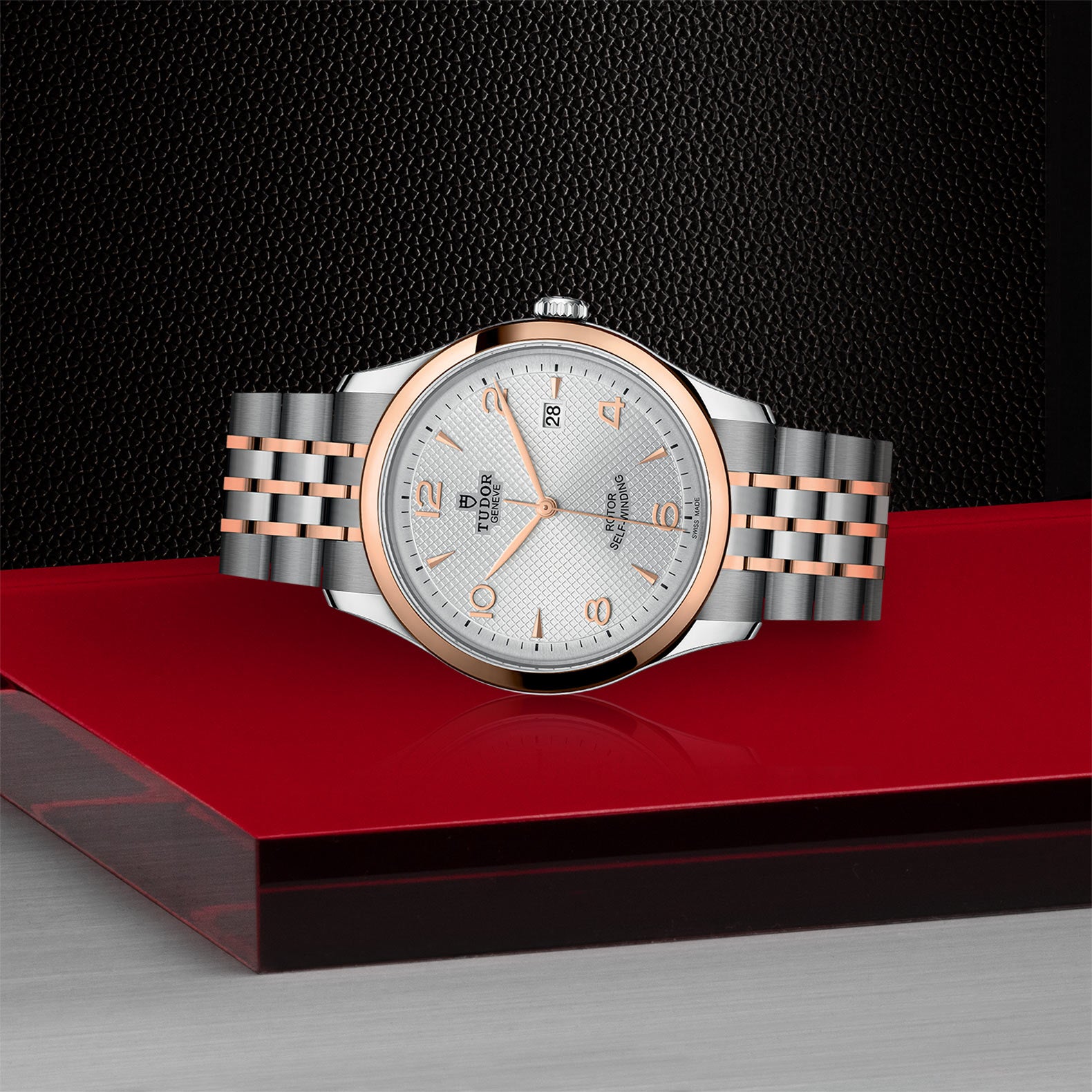 The TUDOR 1926, a two-tone stainless steel and rose gold watch with a silver dial, is shown on a red and black surface with a textured black background—a timeless pick for classic watch enthusiasts.
