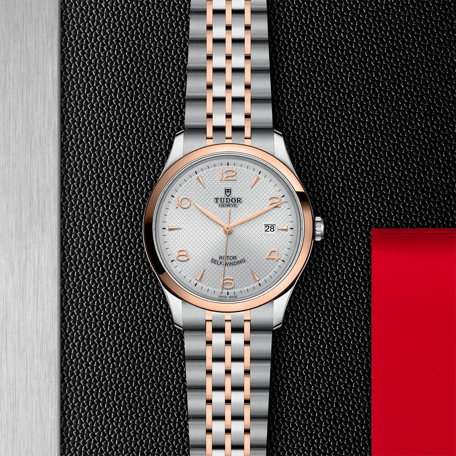 The TUDOR 1926 wristwatch, featuring a silver and rose gold metal band, silver dial, and date display, showcases timeless elegance against a textured black and metallic backdrop with a striking red accent.