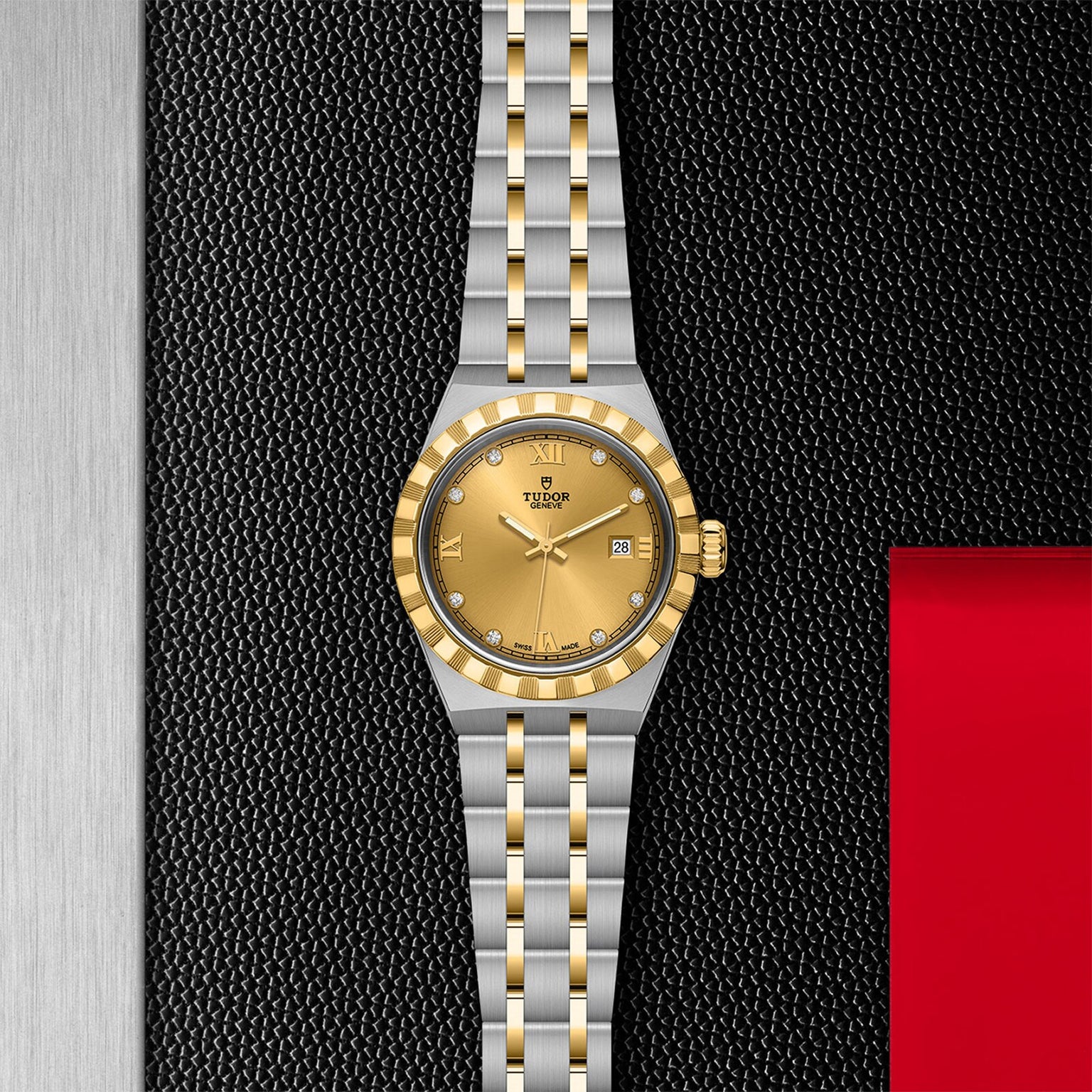 A TUDOR Royal wristwatch with a gold dial, gold bezel, and stainless steel and gold bracelet is displayed vertically on a textured black and silver surface with a red accent.