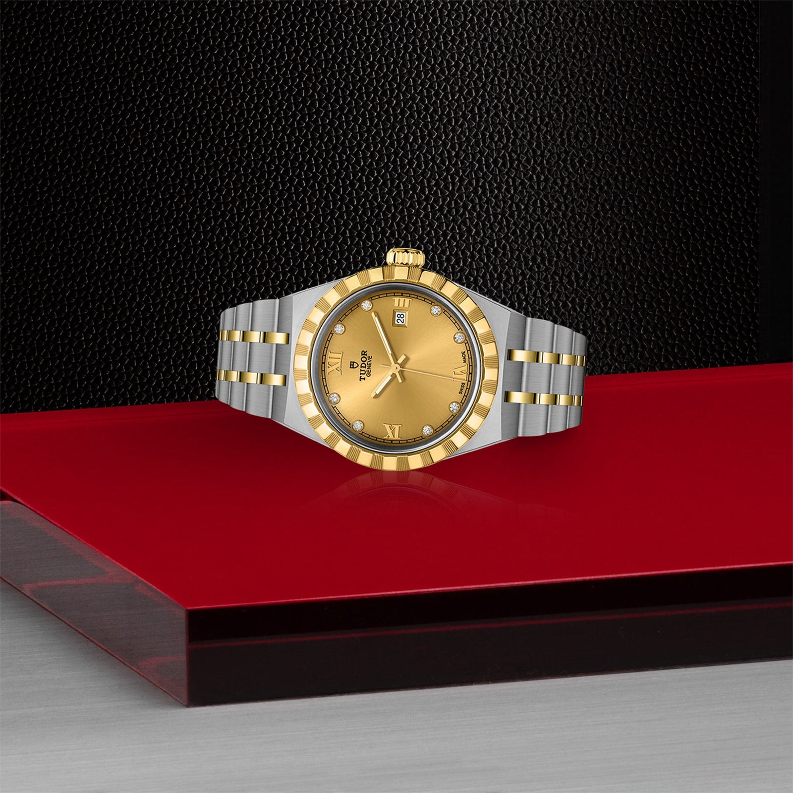 A TUDOR Royal wristwatch in stainless steel and gold with a gold dial and date display, set against a red and black geometric surface and a textured black background.