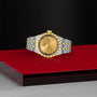 A TUDOR Royal wristwatch in stainless steel and gold with a gold dial and date display, set against a red and black geometric surface and a textured black background.