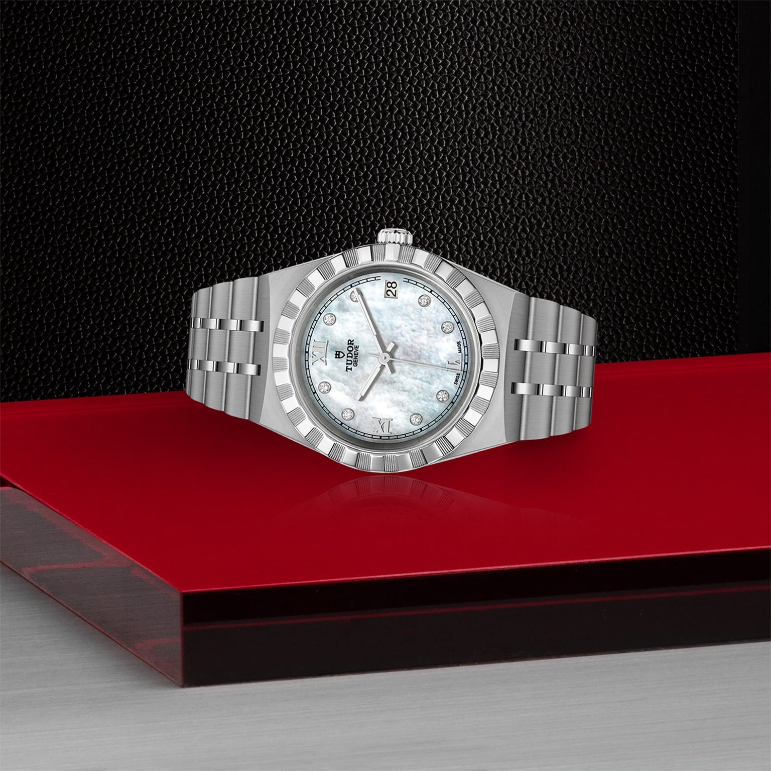 The TUDOR Royal wristwatch, featuring a metallic band and a white mother-of-pearl dial, is showcased on a red glossy platform with a textured black backdrop, exemplifying the sport-chic style of TUDOR watches.