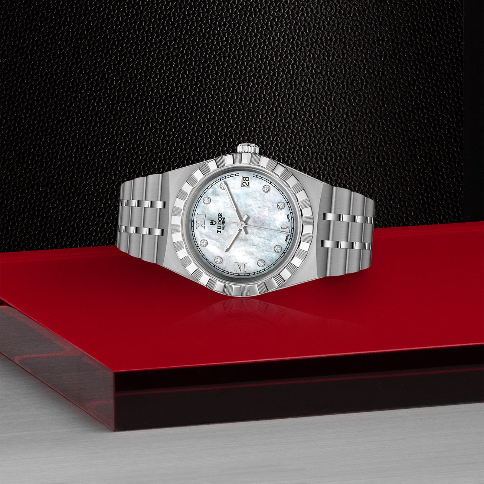 The TUDOR Royal wristwatch, featuring a metallic band and a white mother-of-pearl dial, is showcased on a red glossy platform with a textured black backdrop, exemplifying the sport-chic style of TUDOR watches.