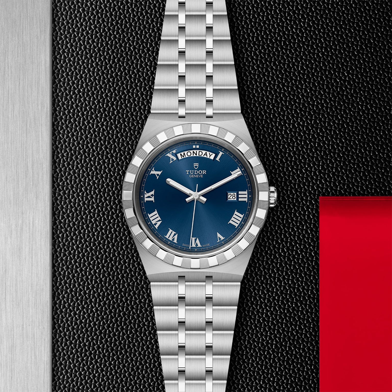 The TUDOR Royal watch features a stainless steel case, blue dial with Roman numerals, day and date display, and an integrated bracelet, displayed against a textured black and red background.